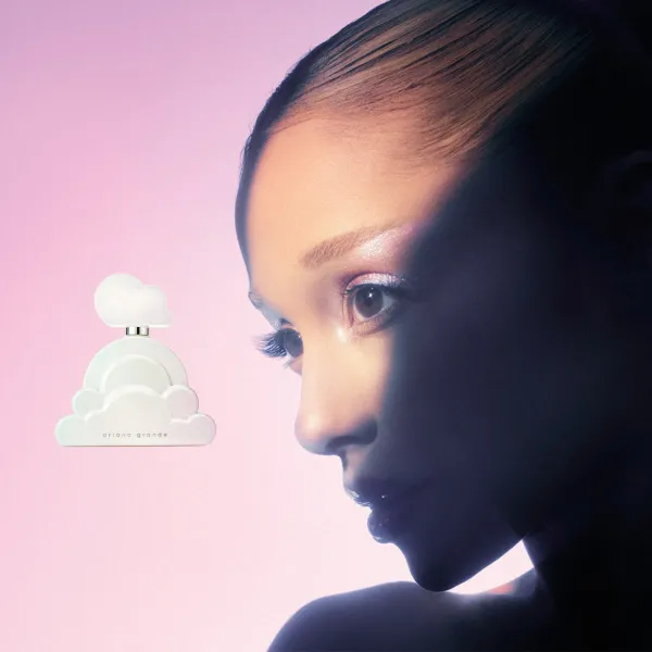 Ariana Grande Cloud smaržu flakons mākoņa formā blakus cilvēka sejas profilam uz maigi rozā fona.