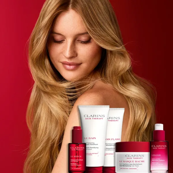 CLARINS Hair Therapy matu kopšanas produktu kolekcija izvietota priekšplānā uz sarkana fona. Aiz produktiem redzama persona ar gariem, viļņainiem matiem, kas izceļ matu spīdumu un kopšanas efektu.