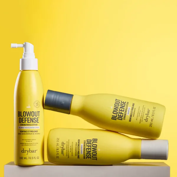 Drybar Blowout Defense matu kopšanas produkti izvietoti uz spilgti dzeltena fona.