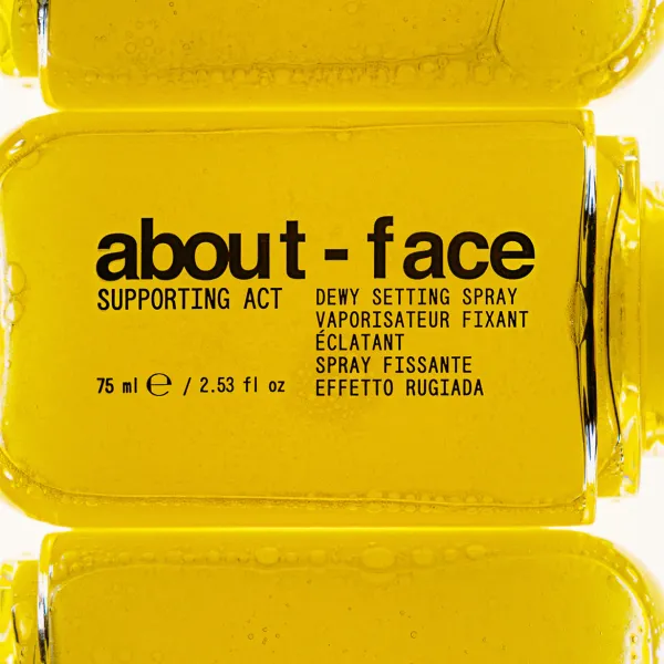 Tuvplāna attēls ar dzeltenām about-face Supporting Act Dewy Setting Spray pudelītēm. Centrā redzams produkta iepakojums ar skaidri salasāmu nosaukumu un tilpumu 75 ml.