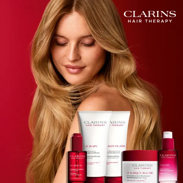 CLARINS Hair Therapy matu kopšanas produktu kolekcija izvietota priekšplānā uz sarkana fona. Aiz produktiem redzama persona ar gariem, viļņainiem matiem, kas izceļ matu spīdumu un kopšanas efektu.