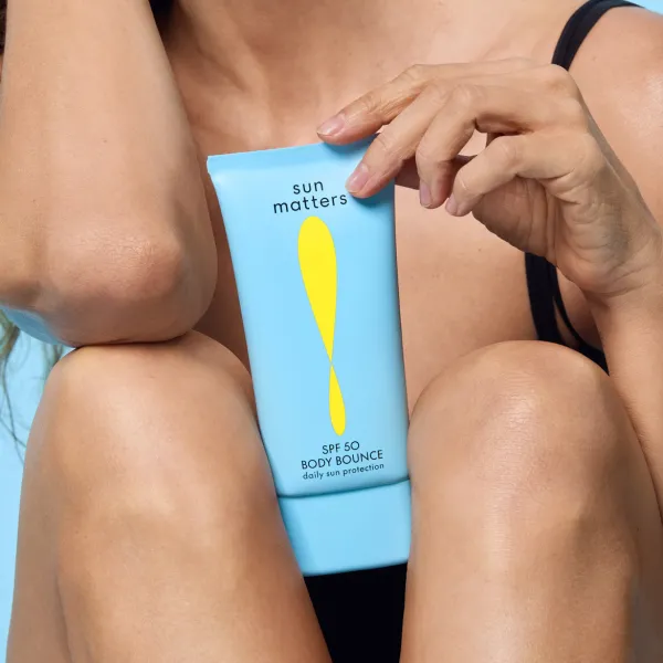 Sieviete tur rokās zilu SPF 50 saules aizsargkrēma tūbiņu „Sun Matters Body Bounce” uz gaiši zila fona.