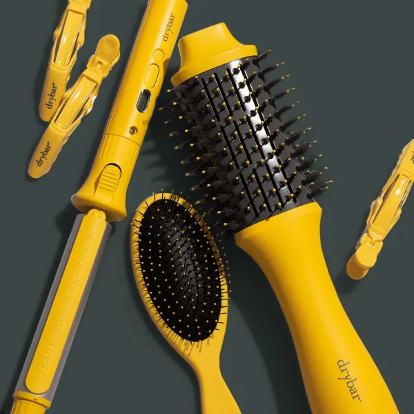 Drybar matu veidošanas rīki un aksesuāri dzeltenā krāsā uz tumši pelēka fona.