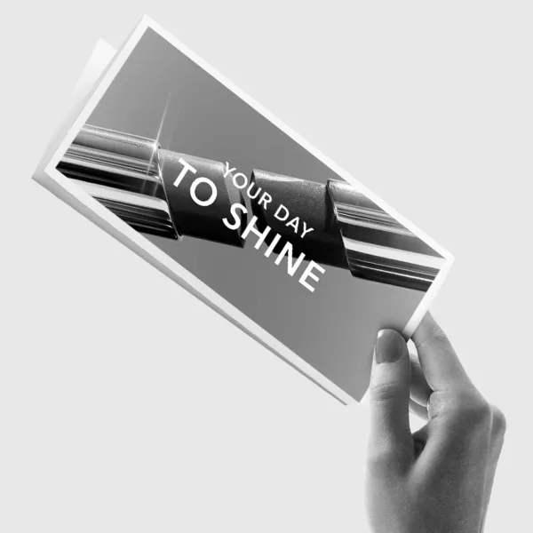 Roka tur atvērtu bukletu ar kosmētikas produkta attēlu un uzrakstu “YOUR DAY TO SHINE” uz gaiša fona.